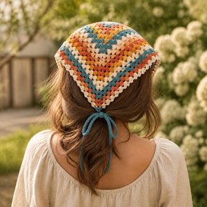 May include: A triangular crochet headscarf featuring a granny square pattern in shades of orange, yellow, teal, and cream. The headscarf is tied at the nape of the neck with two long teal yarn strings that hang down, securing the accessory in place.