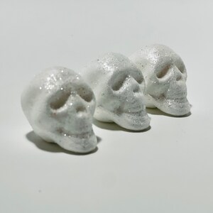 May include: Three white skull figurines covered in glitter. The skulls are arranged in a row, each with detailed facial features. The glitter adds a sparkling effect, enhancing the spooky aesthetic.