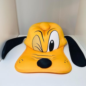 May include: A plush, yellow Pluto-shaped hat with black ears. The hat features Pluto's face, including a winking eye, a black nose, and a smiling mouth. The hat is designed to be worn.