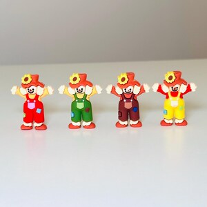 May include: Four colorful clown figurines. Each clown has a red hat with a sunflower, a yellow shirt, and overalls in red, green, brown, and yellow. The clowns have white hands and feet, and a smiling face.