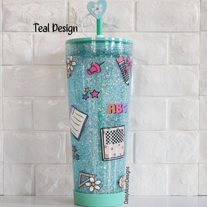 Puede incluir: Un vaso de pl&aacute;stico transparente con purpurina turquesa, una tapa en forma de coraz&oacute;n y una pajita. El vaso est&aacute; decorado con pegatinas de tem&aacute;tica escolar, incluyendo una regla, un l&aacute;piz, una carta, una estrella y un alfiler. El texto "DaisyMoonDesigns" est&aacute; impreso en la parte inferior del vaso.