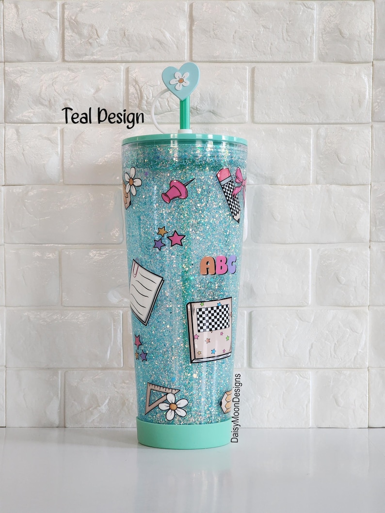 Puede incluir: Un vaso de pl&aacute;stico transparente con purpurina turquesa, una tapa en forma de coraz&oacute;n y una pajita. El vaso est&aacute; decorado con pegatinas de tem&aacute;tica escolar, incluyendo una regla, un l&aacute;piz, una carta, una estrella y un alfiler. El texto "DaisyMoonDesigns" est&aacute; impreso en la parte inferior del vaso.