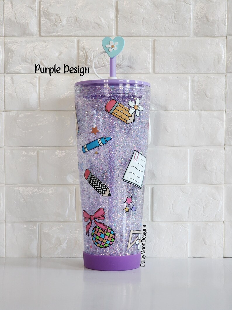Puede incluir: Vaso de purpurina morado con tapa transparente y una pajita. El vaso tiene una variedad de pegatinas con tem&aacute;tica escolar, incluyendo un l&aacute;piz, un cray&oacute;n, una regla, un cuaderno y un lazo. El texto "DaisyMoonDesigns" es visible en el vaso.