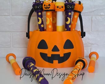 Puede incluir: Un cubo de calabaza de plástico naranja lleno de varios bolígrafos cilíndricos con adornos de Halloween, incluyendo gatos negros con sombreros morados, calabazas naranjas y dulces de maíz. Otros bolígrafos yacen en la superficie frontal.