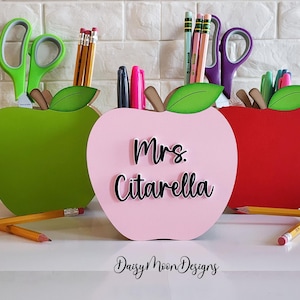 May include: Three wooden apple-shaped desk organizers, one green, one pink with "Mrs. Citarella" written on it, and one red. Each organizer has a slot for pens, pencils, and scissors.