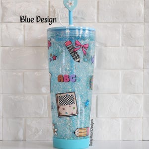 Puede incluir: Un vaso azul con purpurina, tapa transparente y pajita. El vaso tiene una variedad de pegatinas coloridas con tem&aacute;tica escolar, incluyendo un l&aacute;piz, un libro y las letras "ABC". El texto "DaisyMoonDesigns" est&aacute; impreso en el lateral del vaso.