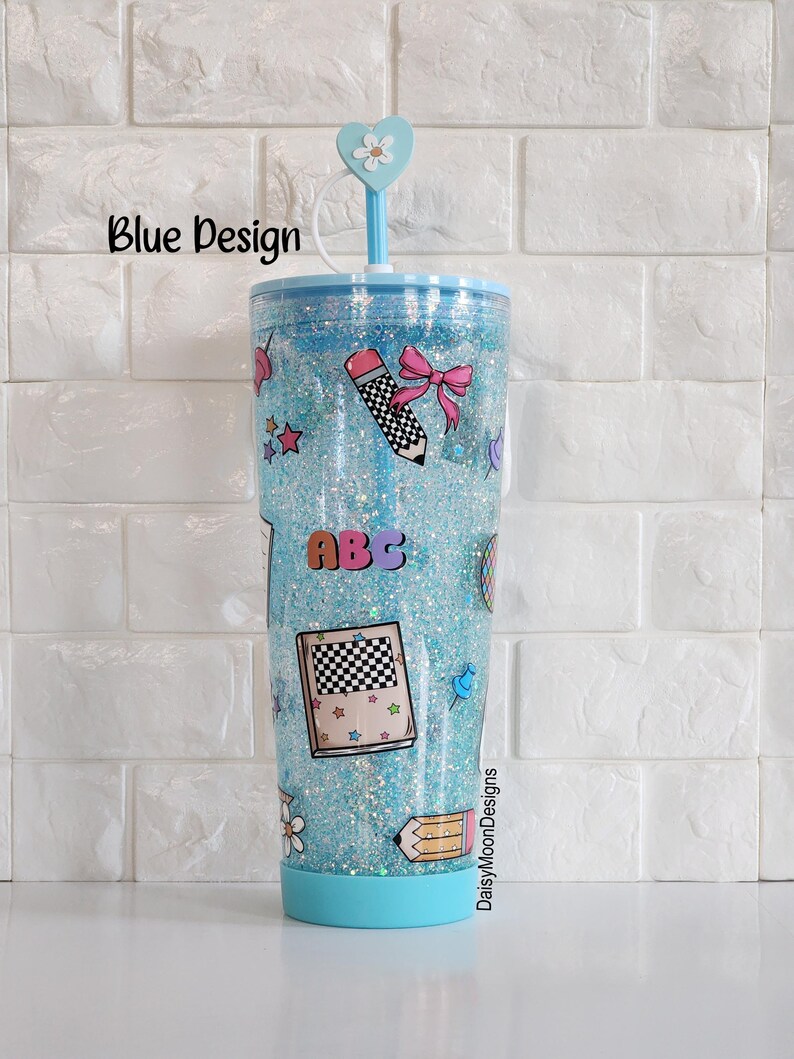 Puede incluir: Un vaso azul con purpurina, tapa transparente y pajita. El vaso tiene una variedad de pegatinas coloridas con tem&aacute;tica escolar, incluyendo un l&aacute;piz, un libro y las letras "ABC". El texto "DaisyMoonDesigns" est&aacute; impreso en el lateral del vaso.
