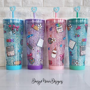 Puede incluir: Tres vasos de glitter con tem&aacute;tica de regreso a clases. Los vasos son azul, morado y rosa con glitter y pegatinas con tem&aacute;tica escolar. Los vasos tienen tapas transparentes y pajitas.