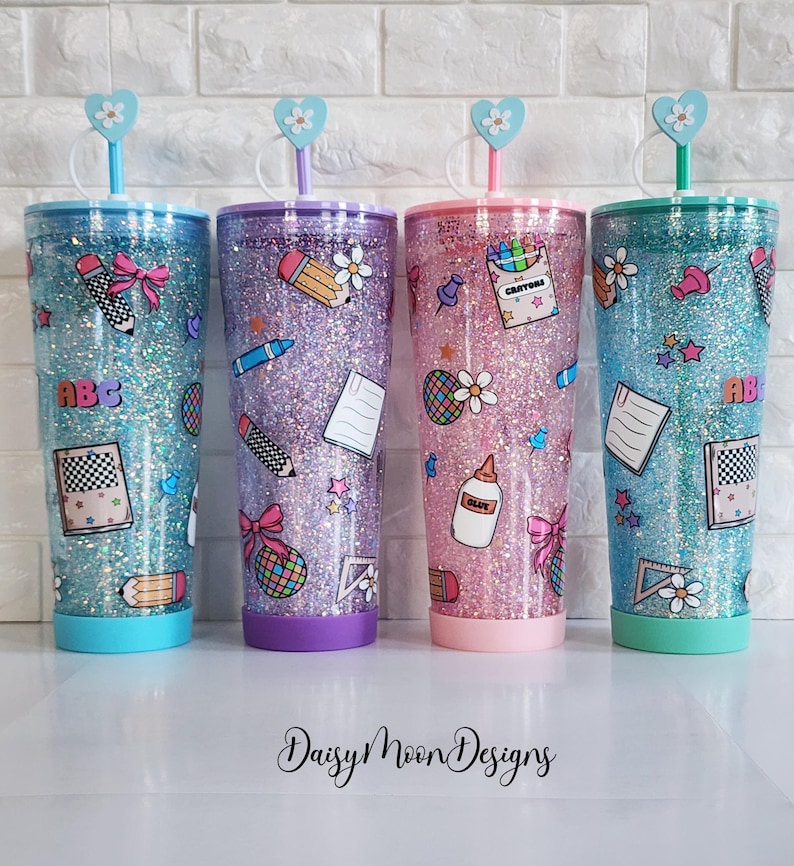 Puede incluir: Tres vasos de glitter con tem&aacute;tica de regreso a clases. Los vasos son azul, morado y rosa con glitter y pegatinas con tem&aacute;tica escolar. Los vasos tienen tapas transparentes y pajitas.