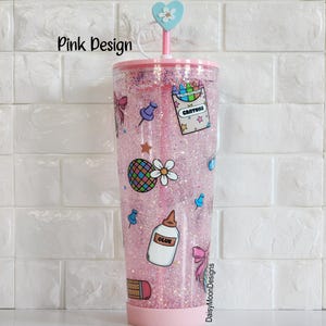 Puede incluir: Un vaso rosa con purpurina con una tapa en forma de coraz&oacute;n y una pajita. El vaso presenta una variedad de ilustraciones con tem&aacute;tica escolar, incluyendo l&aacute;pices de colores, un l&aacute;piz, pegamento, chinchetas y una margarita. El texto "CRAYONS" y "GLUE" es visible en el vaso.