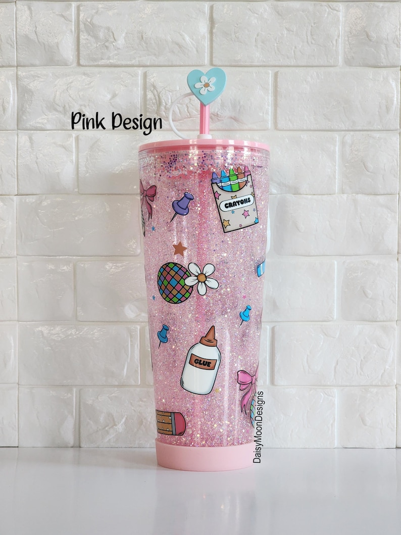 Puede incluir: Un vaso rosa con purpurina con una tapa en forma de coraz&oacute;n y una pajita. El vaso presenta una variedad de ilustraciones con tem&aacute;tica escolar, incluyendo l&aacute;pices de colores, un l&aacute;piz, pegamento, chinchetas y una margarita. El texto "CRAYONS" y "GLUE" es visible en el vaso.