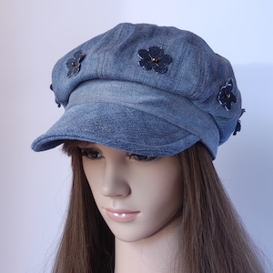 Op de afbeelding: Een denim newsboy-pet met een afgeronde kroon en een korte, gebogen klep. De blauwe pet is versierd met verschillende donkerblauwe bloemenapplicaties. De pet wordt getoond op een mannequin met lang bruin haar.
