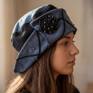 May include: A blue and black beret-style hat with a decorative black flower and silver studs. The hat has a geometric design with black stitching and is made of a soft, suede-like material. The hat is a stylish accessory.