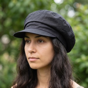 Op de afbeelding: Een zwarte baret met een korte klep. De pet is gemaakt van een zachte, getextureerde stof en heeft een ronde kroon. De hoed wordt gedragen door een persoon met lang, donker haar.