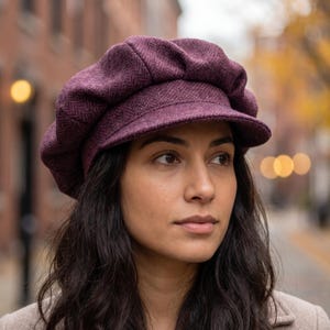 Pode incluir: Uma boina de estilo newsboy em roxo escuro com um padrão subtil de espinha de peixe. O chapéu tem uma coroa estruturada e arredondada com dobras suaves e uma pala curta e rígida na frente, feita de um tecido texturizado com aspeto tecido.