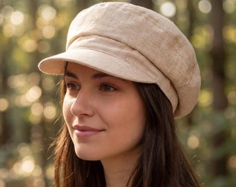 May include: A beige newsboy cap with a short brim. The cap has a textured, plaid-like pattern and a rounded crown. The hat is made of a soft material and is suitable for various occasions. The background is a blurred forest scene.