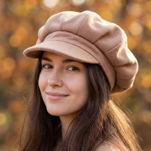 Può includere: Un cappello newsboy color beige con una corona arrotondata e una visiera corta e rigida. Il cappello è realizzato in un materiale morbido e strutturato. La persona ha lunghi capelli castano scuro.