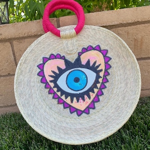 May include: A round straw bag with a pink and white heart-shaped design featuring a blue evil eye. The bag has a pink handle.