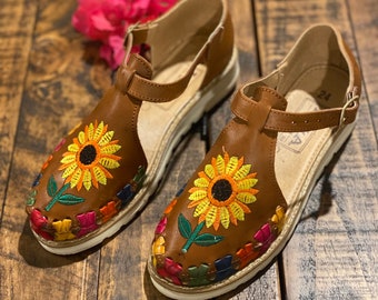 May include: A pair of brown leather sandals with embroidered sunflowers and colorful accents. The sandals have a white platform sole and a strap across the ankle.