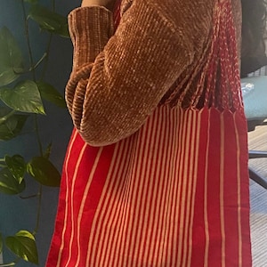 Puede incluir: Bolso tote a rayas rojas y beige con un diseño tejido. El bolso se lleva al hombro, mostrando sus rayas verticales. Las correas del bolso son rojas y tienen un detalle de flecos. La persona lleva un suéter marrón.
