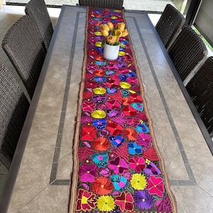 Peut inclure: Un chemin de table brodé coloré avec un motif floral dans des tons de rose, rouge, jaune, bleu et vert. Le chemin de table a une bordure à franges et est présenté sur une table avec des chaises en osier.