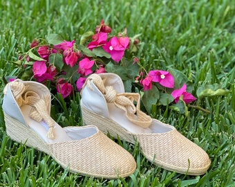 Könnte beinhalten: Ein Paar beige Espadrillesandalen mit gewebten Riemen und einem Keilabsatz. Die Sandalen liegen auf einem Bett aus grünem Gras mit rosa Blumen.