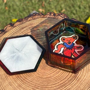 May include: A hexagonal resin box with a lid. The box is open, revealing a colorful interior and a sticker of a dancing bear. The lid is white with a silver shimmer. The box sits on a wooden surface.