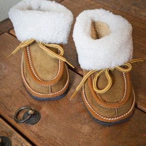 Puede incluir: Un par de zapatillas de ante marrón con forro de piel sintética blanca y cordones amarillos. Las zapatillas están sobre una superficie de madera.