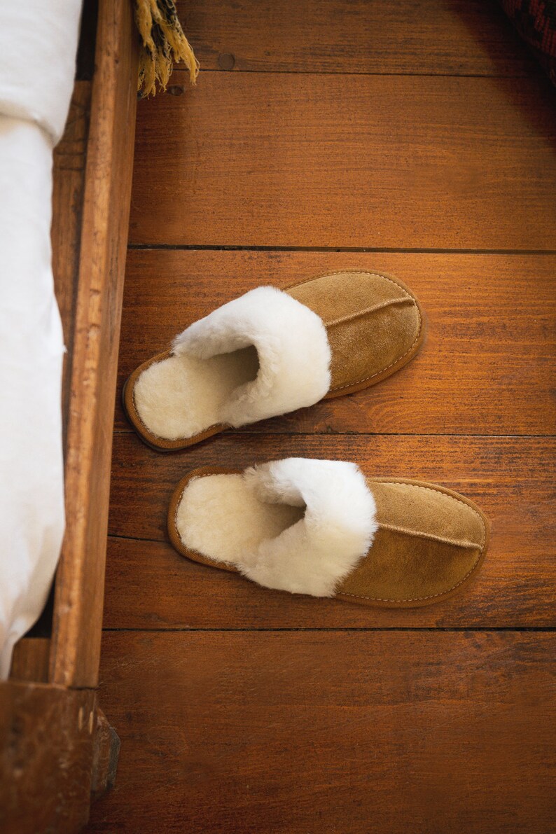 May include: A pair of brown suede slippers with white faux fur lining. The slippers are on a wooden floor.