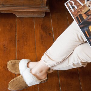 May include: A pair of brown suede slippers with white faux fur lining. The slippers are worn by a person sitting on a wooden floor.