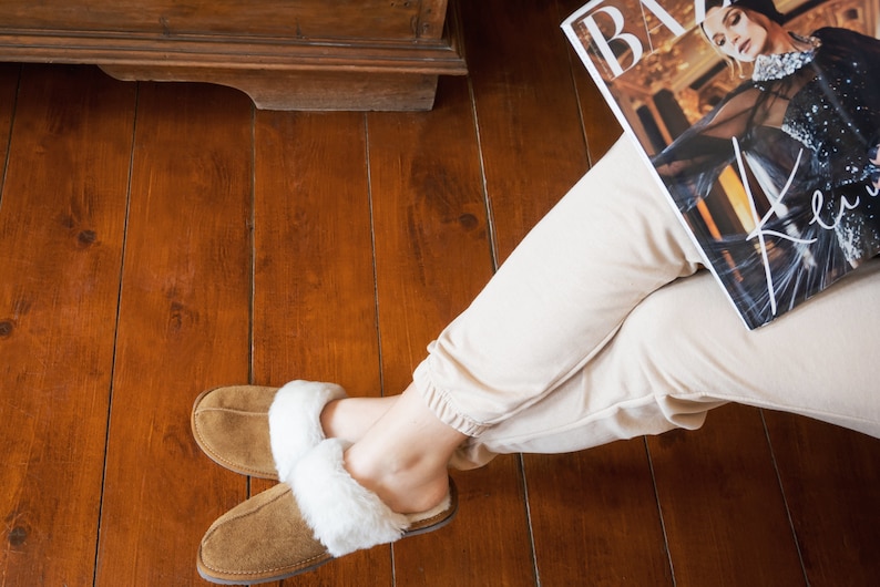 May include: A pair of brown suede slippers with white faux fur lining. The slippers are worn by a person sitting on a wooden floor.