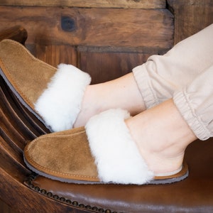 May include: A pair of brown suede slippers with white faux fur trim. The slippers are worn by a person sitting on a wooden chair.