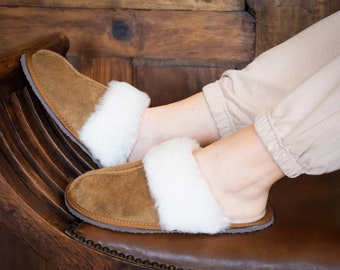 May include: A pair of brown suede slippers with white faux fur trim. The slippers are worn by a person sitting on a wooden chair.