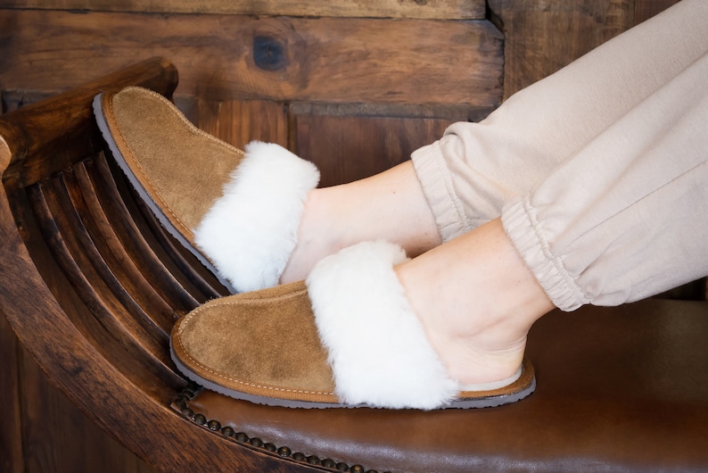 May include: A pair of brown suede slippers with white faux fur trim. The slippers are worn by a person sitting on a wooden chair.