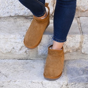 Peut inclure: Une paire de bottines en daim marron avec une doublure en peau de mouton blanche. Les bottes sont portées par une personne portant un jean bleu.