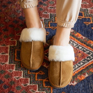 May include: A pair of brown suede slippers with white faux fur trim. The slippers are on a colorful rug.