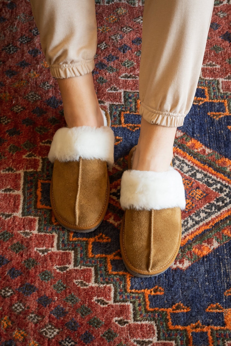 May include: A pair of brown suede slippers with white faux fur trim. The slippers are on a colorful rug.
