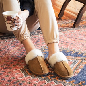 May include: A pair of brown suede slippers with white faux fur trim. The slippers are worn by a person sitting on a patterned rug.