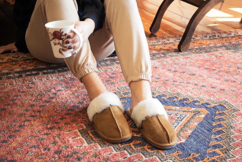 May include: A pair of brown suede slippers with white faux fur trim. The slippers are worn by a person sitting on a patterned rug.
