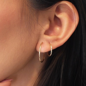 Puede incluir: Primer plano de una oreja con dos pendientes de aro dorados. Los pendientes son de diferentes tamaños, uno es más pequeño que el otro. La persona tiene el pelo largo y oscuro y se toca la cara con la mano.