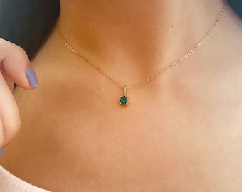 May include: A delicate gold necklace featuring a small, square-cut, dark green gemstone pendant. The pendant is suspended from a thin gold chain, creating a minimalist and elegant design. The necklace is worn around the neck.
