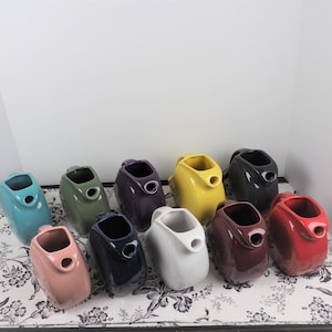 May include: A collection of 10 ceramic creamer pitchers in various colors, including teal, green, purple, yellow, white, red, and blue. The pitchers are arranged in a row on a white surface with a floral pattern.