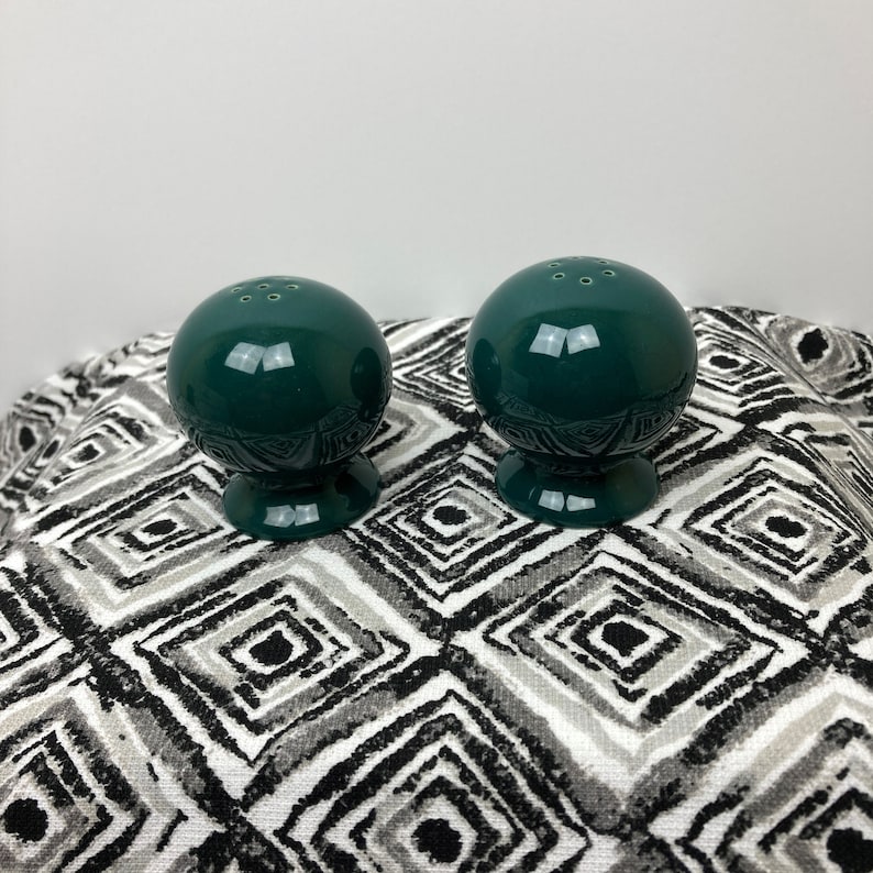 May include: A pair of green ceramic salt and pepper shakers with a round shape and a small base. The shakers have a glossy finish and are sitting on a black and white patterned surface.