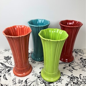May include: Four ceramic vases with a fluted, scalloped rim and vertical ribbed detailing. The vases are finished in glossy solid colors: coral, teal, lime green, and red. Each vase features a flared opening and a circular base.