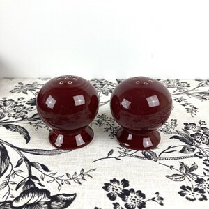 May include: Two red ceramic salt and pepper shakers with a round shape and a small hole on top for dispensing salt and pepper.