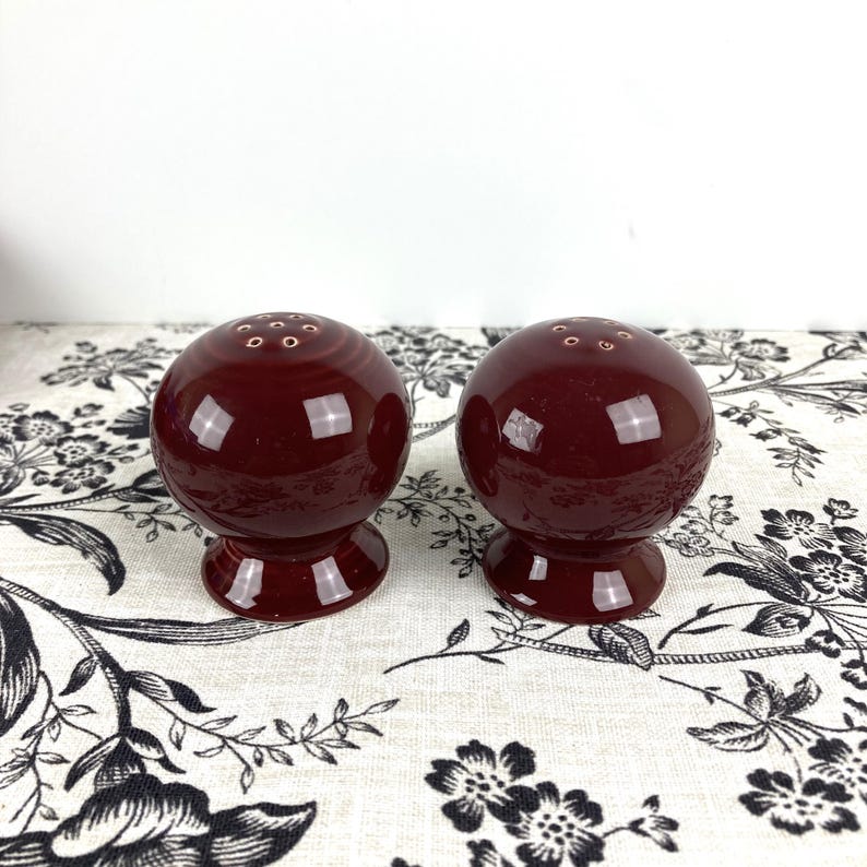 May include: Two red ceramic salt and pepper shakers with a round shape and a small hole on top for dispensing salt and pepper.