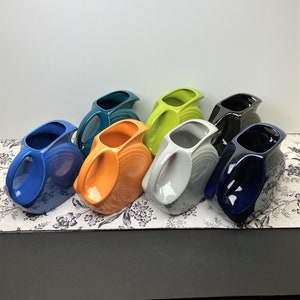 May include: A set of eight ceramic pitchers in various colors, including blue, green, orange, gray, and black. The pitchers have a unique, modern design with a curved handle and a wide spout.