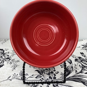 Peut inclure: Un bol rond en c&eacute;ramique rouge profond avec une finition brillante. L'int&eacute;rieur du bol pr&eacute;sente une s&eacute;rie d'anneaux circulaires concentriques en relief sur la base. Le bol est soutenu par un support de pr&eacute;sentation en fil m&eacute;tallique noir torsad&eacute;.
