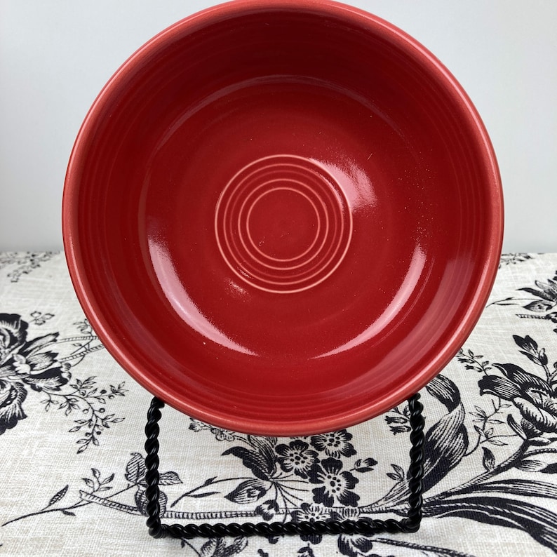 Peut inclure: Un bol rond en c&eacute;ramique rouge profond avec une finition brillante. L'int&eacute;rieur du bol pr&eacute;sente une s&eacute;rie d'anneaux circulaires concentriques en relief sur la base. Le bol est soutenu par un support de pr&eacute;sentation en fil m&eacute;tallique noir torsad&eacute;.