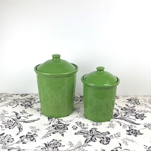May include: Two green ceramic canisters with lids. The larger canister is taller and wider than the smaller canister. Both canisters have a smooth, glossy finish.
